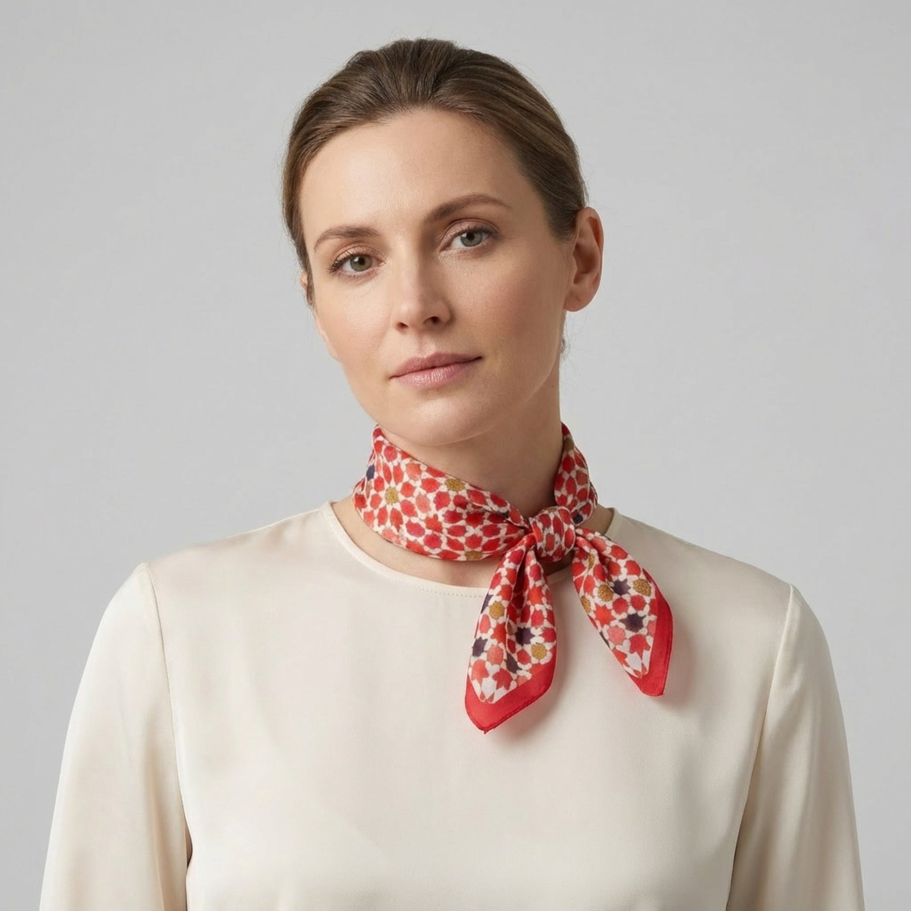 A woman with light brown hair wears a cream blouse and the Small Square Scarf Alhambra, featuring a red pattern, tied around her neck against a plain gray background.