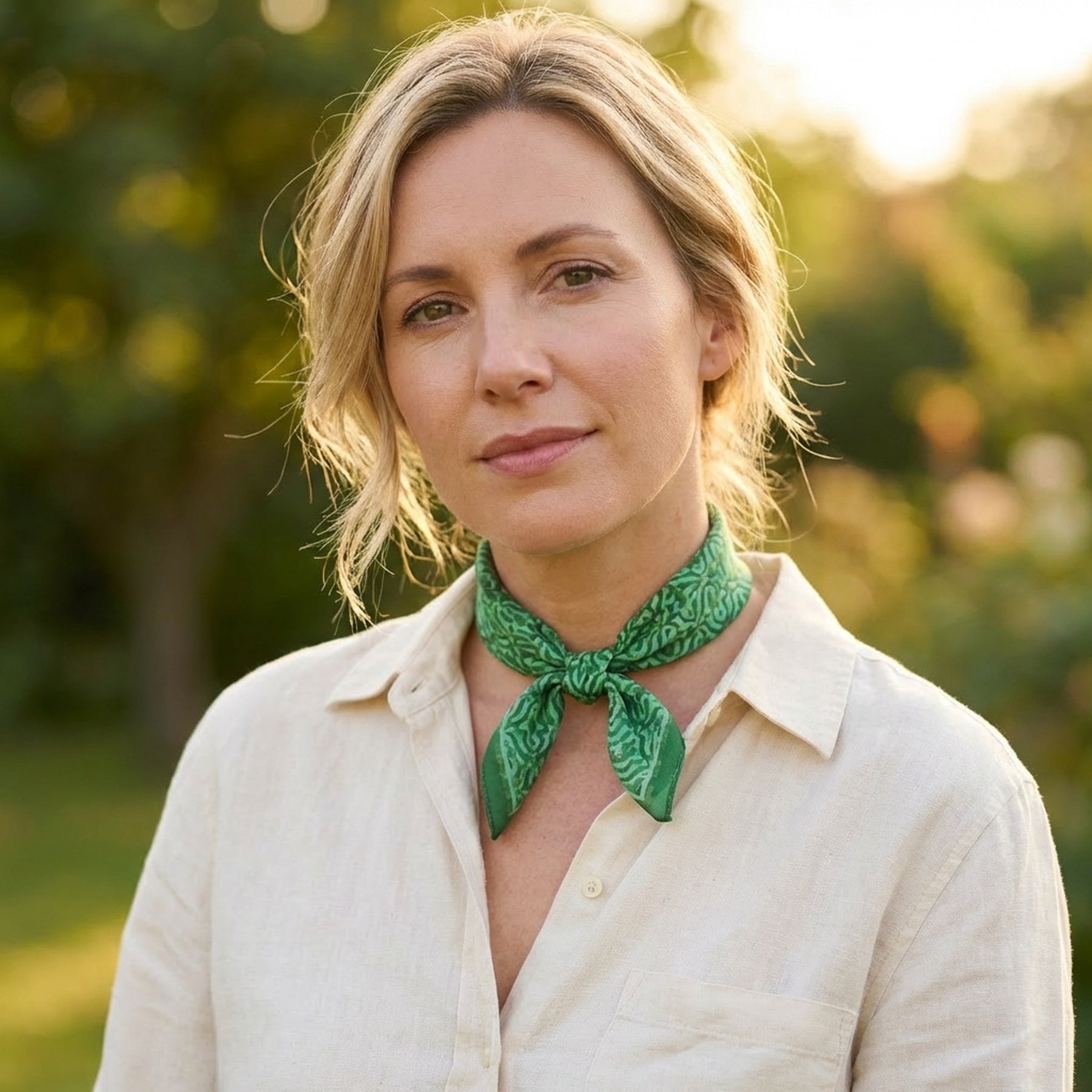 A woman with blonde hair, in a light shirt and the Green Square Silk Scarf Fihri featuring an Islamic art print, stands outdoors amid greenery in soft sunlight.