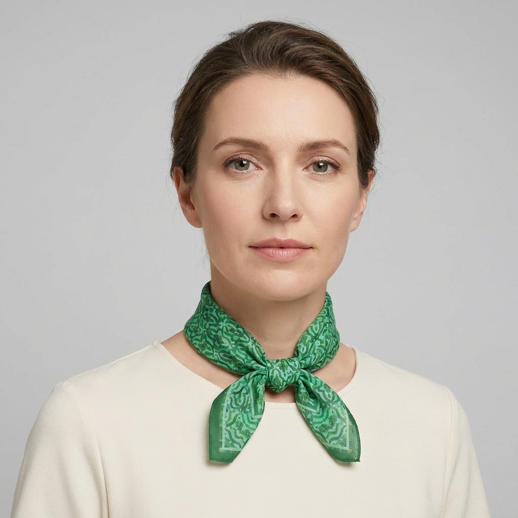 A woman with light brown hair tied back, in a cream top and the Green Square Silk Scarf Fihri featuring an Islamic art print, poses against a plain light gray background.