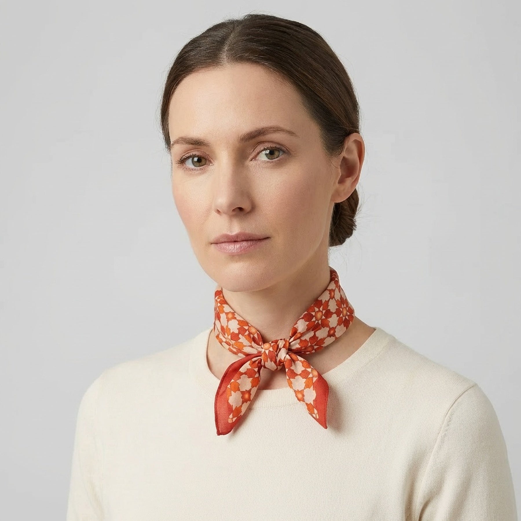 A woman with light skin and brown hair pulled back wears a cream top and the Silk Neckerchief Orange Islamic Art Print, standing against a plain light gray background.