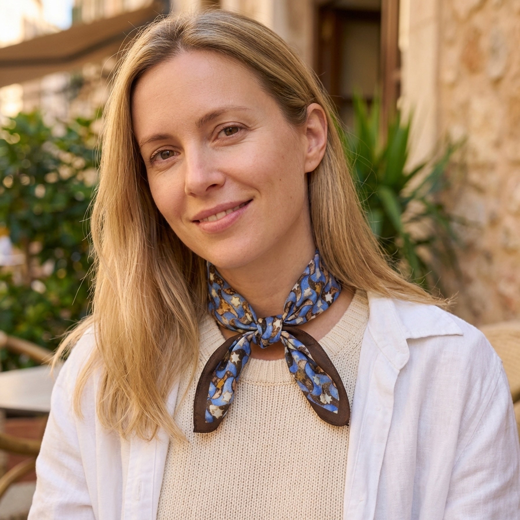 A woman with long blonde hair sits outdoors, smiling, in a cream sweater, white shirt, and the Silk Square Scarf for Women Brown featuring an Islamic art print tied around her neck. Green plants and a stone building are in the background.