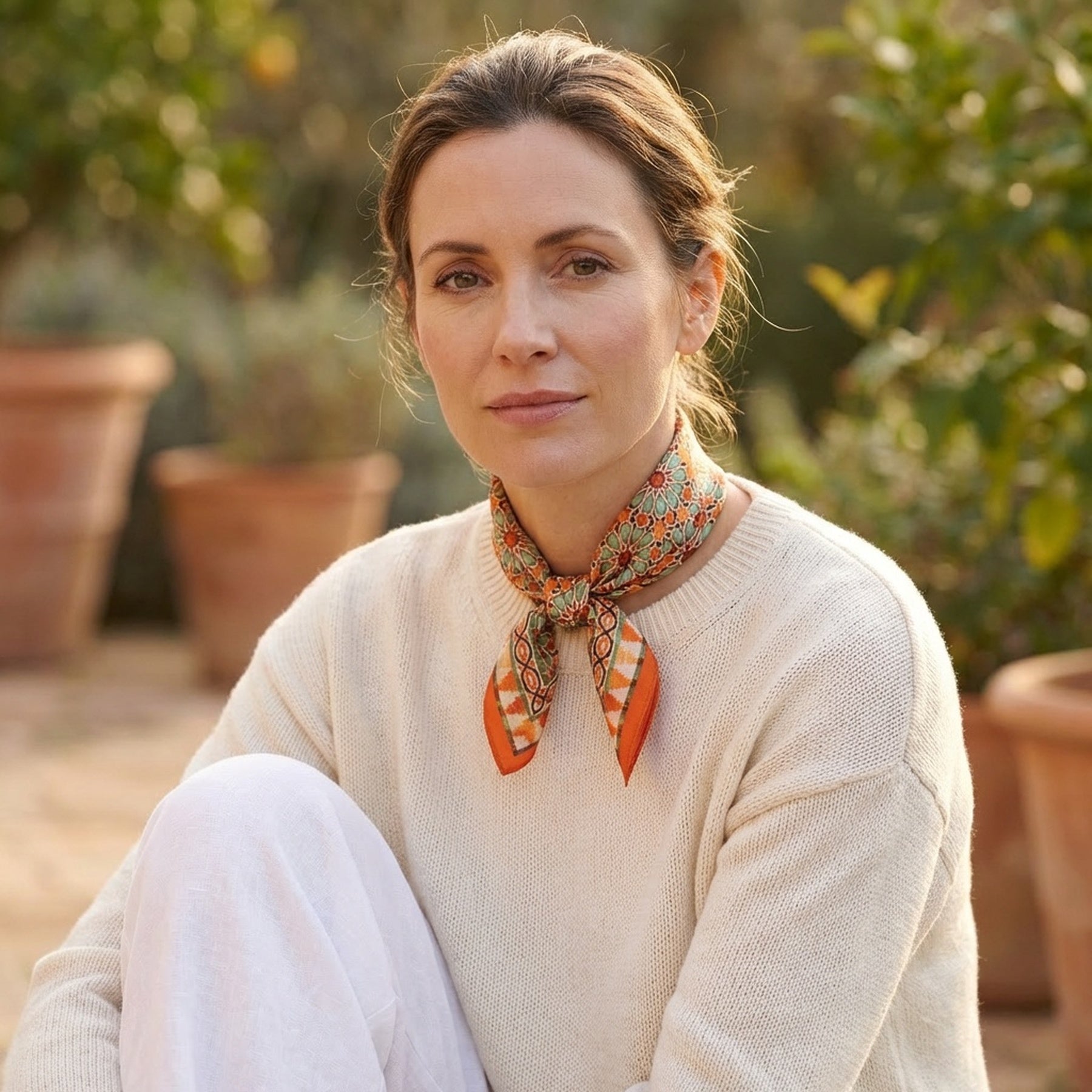 A woman with light brown hair tied back wears a cream sweater, white pants, and the Orange Square Scarf Zellige around her neck while sitting outdoors with potted plants in the background.