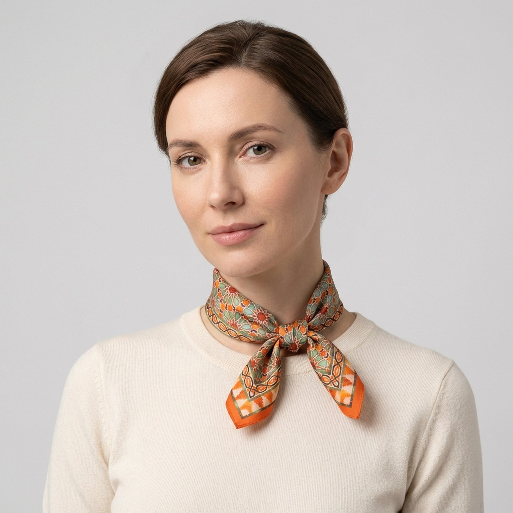 A woman with light skin and brown hair wears a cream-colored top and the Orange Square Scarf Zellige tied around her neck, standing against a plain light gray background.