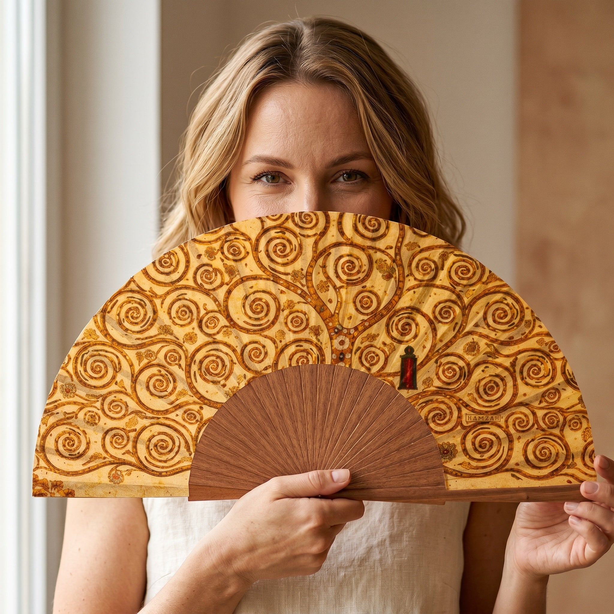 A woman holding the Gustav Klimt Print Hand Fan Tree of Life, with its Gustav Klimt Tree of Life golden print, against a soft neutral background.