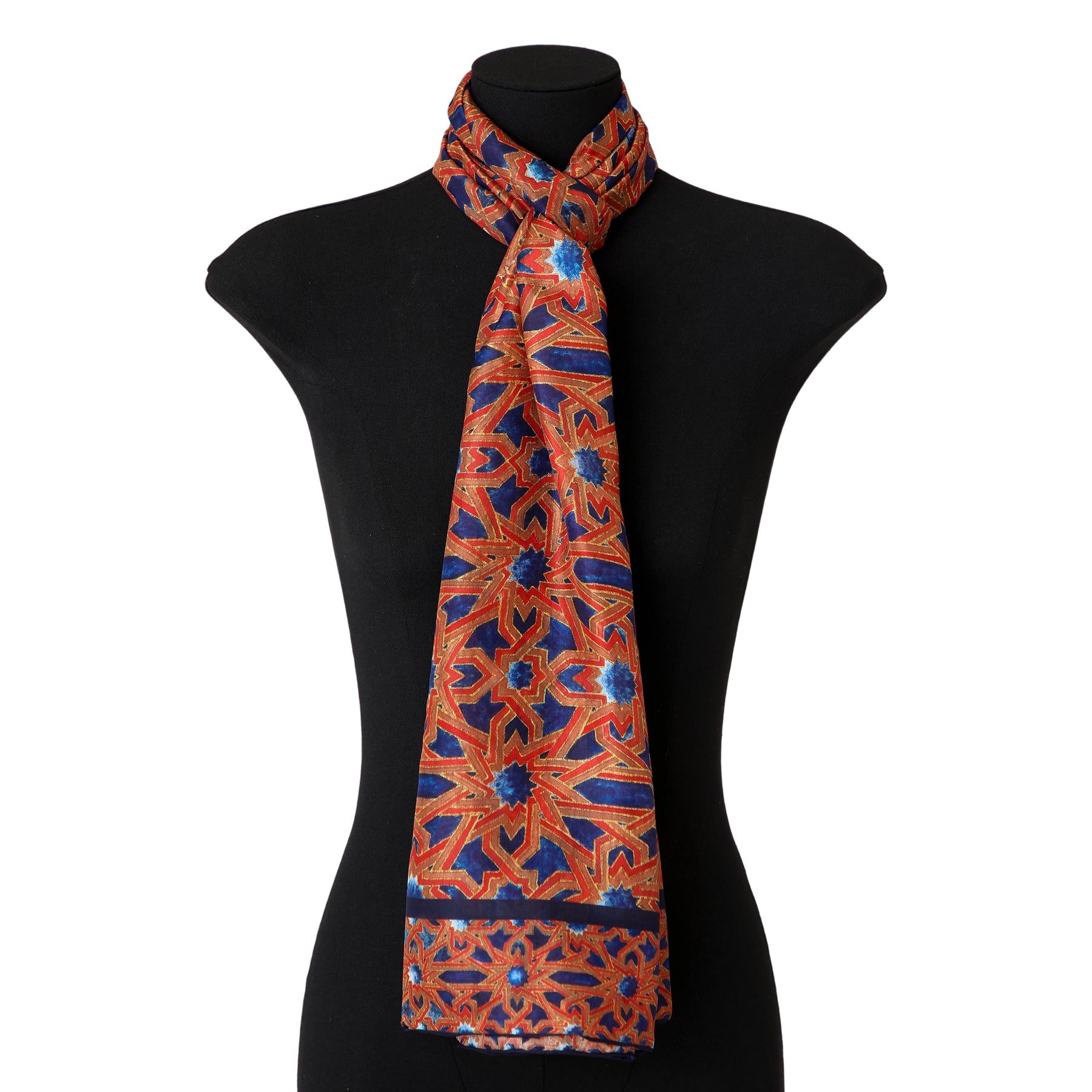 A black mannequin displays the Islamic Art Silk Scarf Alcázar Genil, featuring long silk fabric with vibrant red, blue, and gold geometric patterns tied loosely around the neck.