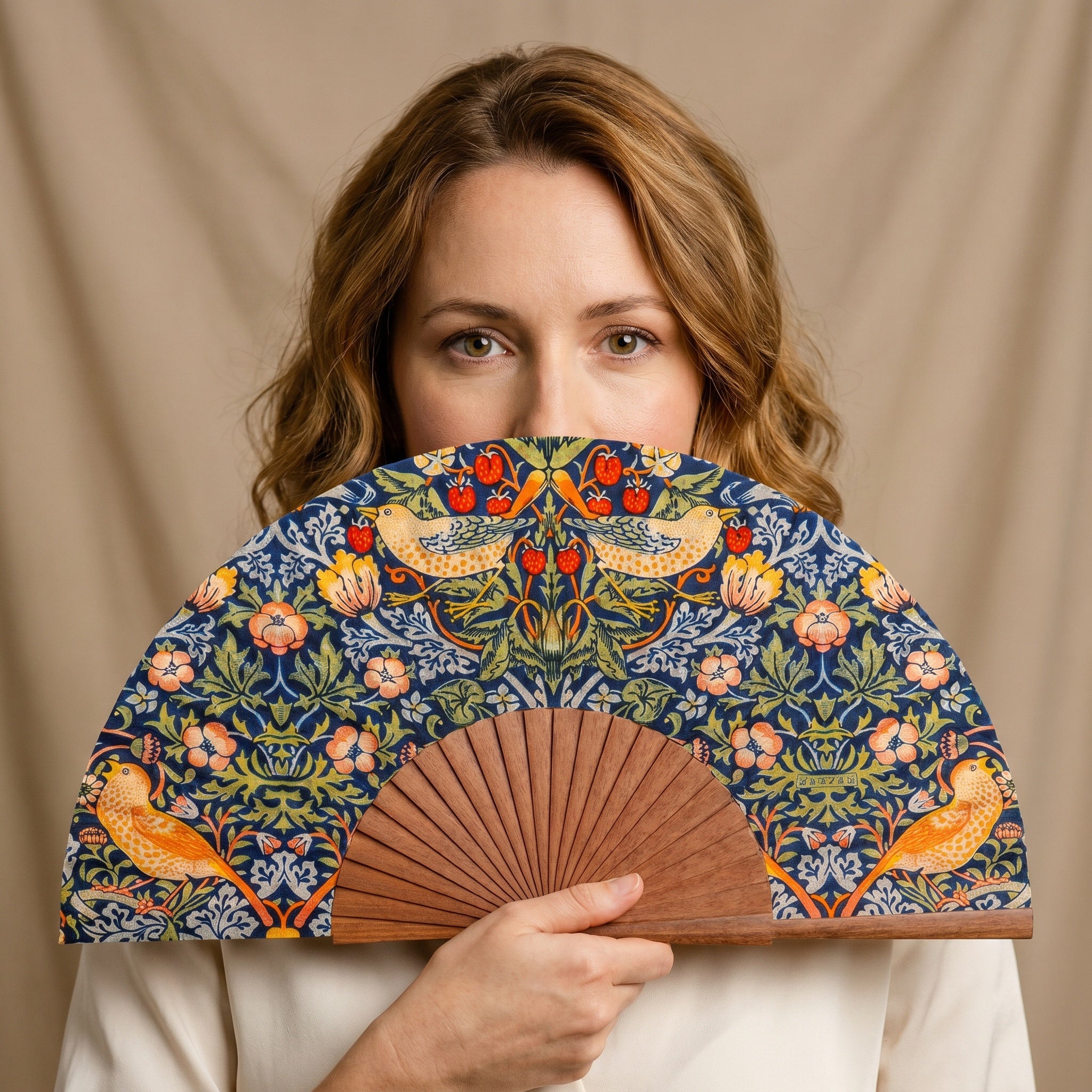 A woman holding the William Morris Print Hand Fan Birds II, with its William Morris Strawberry Thief print on dark silk, against a soft neutral background.