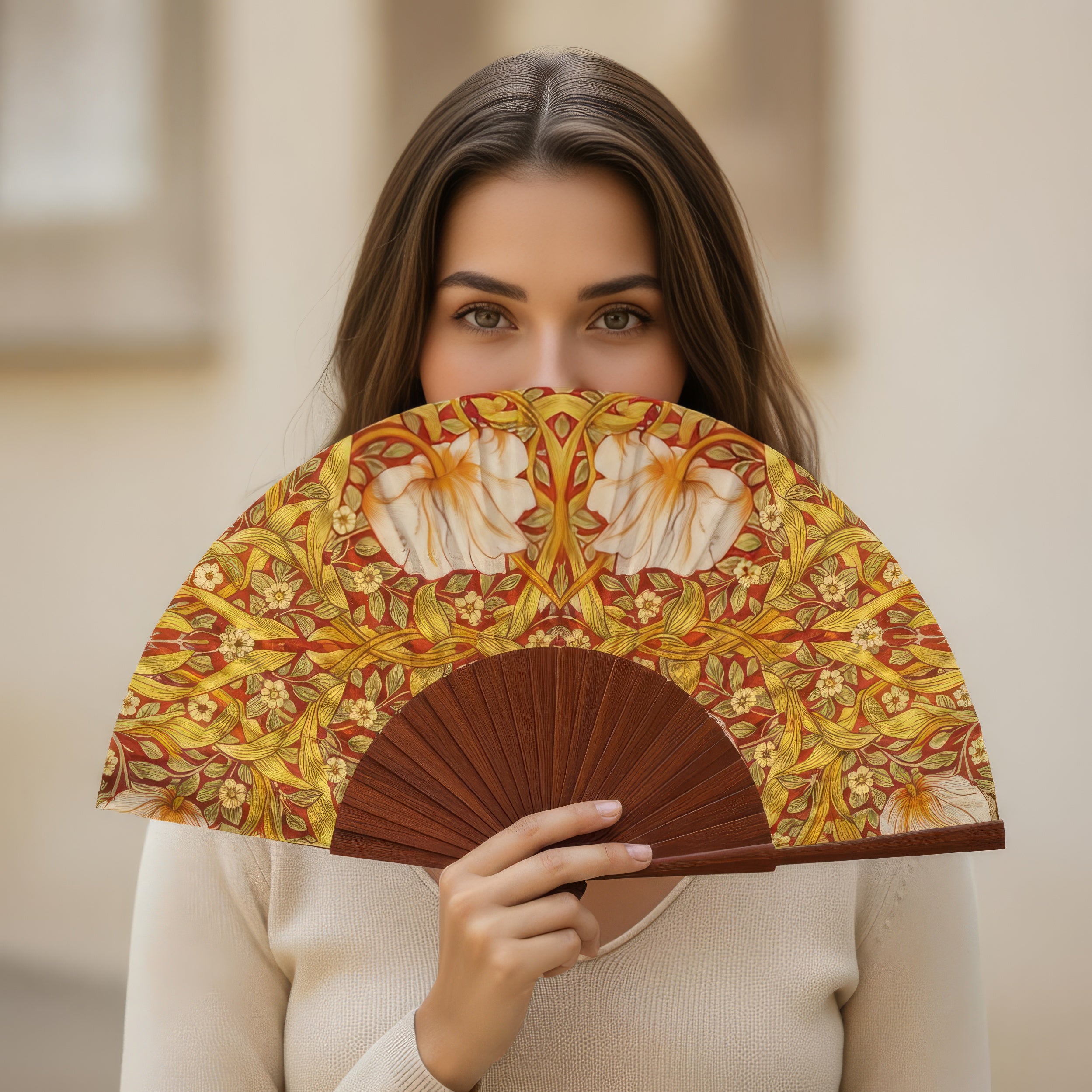 A woman with long brown hair, wearing a light sweater and standing outdoors, partially covers her face with the Hand Fan Made in Spain Basile—its warm yellow and orange tones evoke a William Morris print.