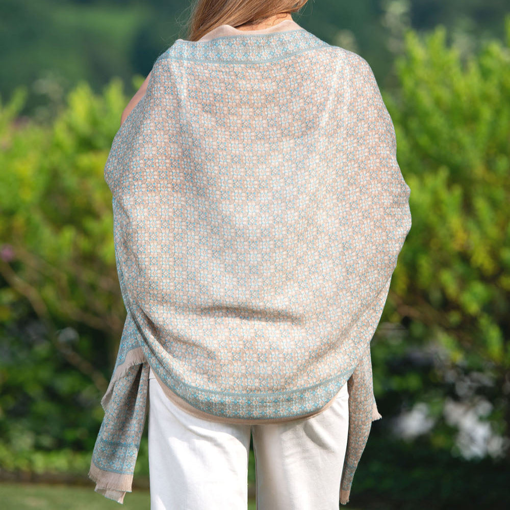 Back view of a model showcasing a beige and blue silk scarf with an intricate mosaic pattern. The lightweight fabric elegantly drapes over the shoulders, highlighting its soft texture and delicate craftsmanship.