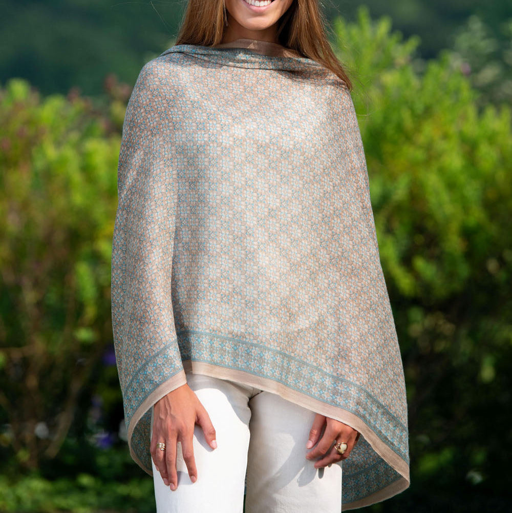 Model wearing a beige and blue silk scarf draped in a poncho-style, featuring an elegant mosaic pattern. The soft, lightweight fabric flows effortlessly, offering a refined and sophisticated look.