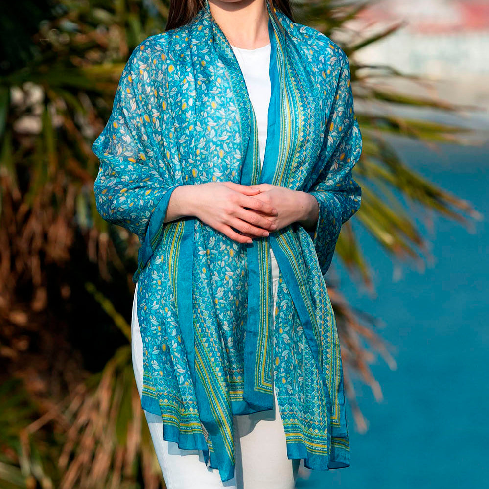 Elegant blue silk scarf with intricate floral print, draped over a model by the seaside.