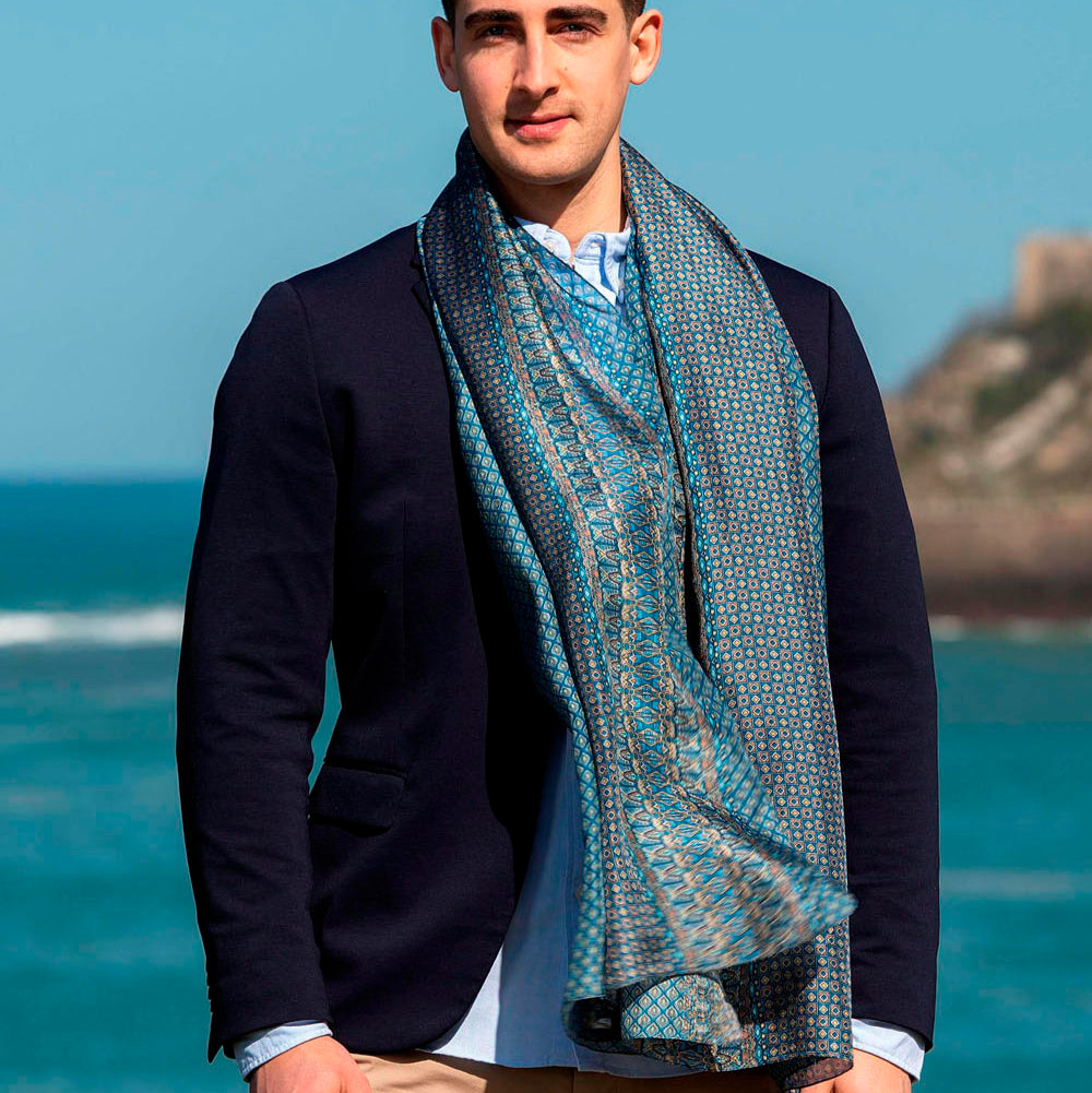 Man wearing a luxurious blue silk scarf with intricate geometric patterns, elegantly draped over a navy suit, standing by the seaside.