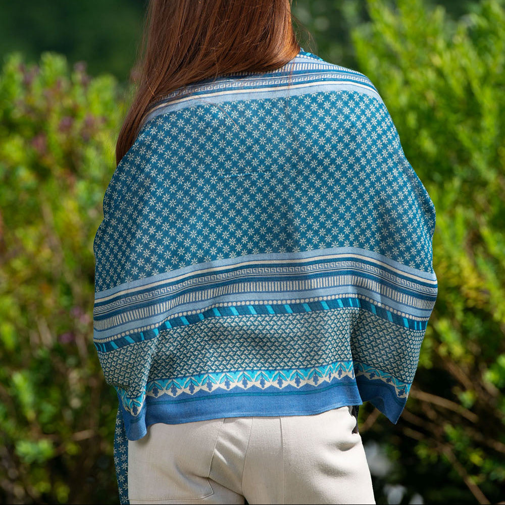 Model seen from the back wearing a blue wool and silk scarf with an intricate geometric pattern. The lightweight fabric elegantly drapes over the shoulders, showcasing delicate details and a sophisticated design.