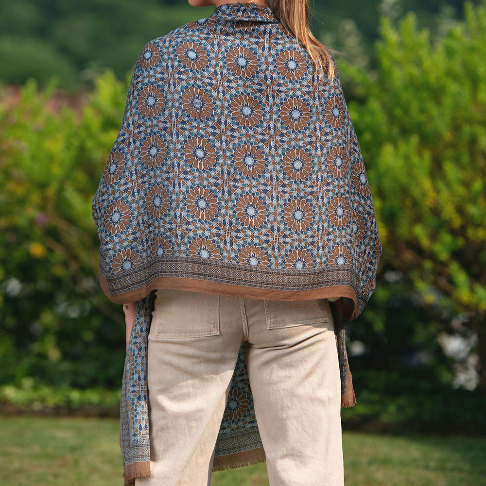 Model wearing a luxurious silk scarf featuring an intricate mosaic pattern in earthy brown and blue tones. The geometric design, inspired by traditional craftsmanship, drapes elegantly over the shoulders, complementing a natural outdoor setting.