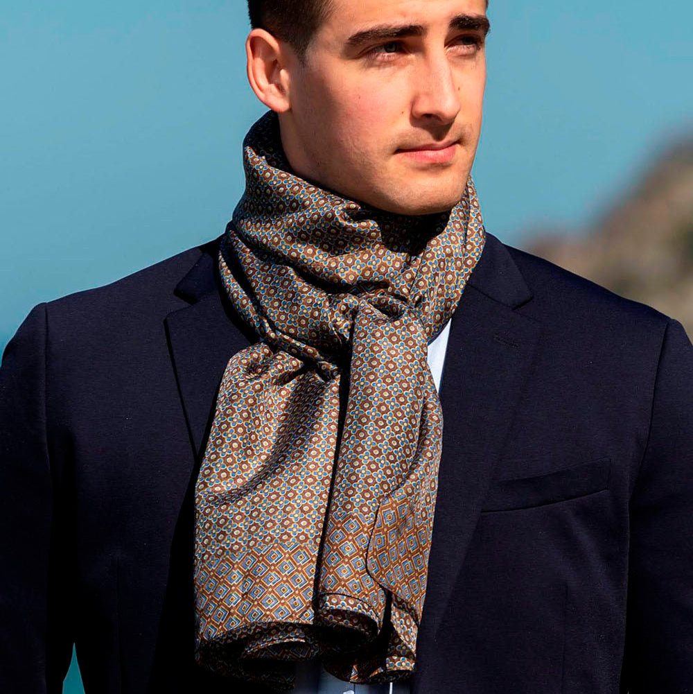 Man wearing a sophisticated brown silk scarf with an intricate geometric pattern in warm earthy tones, elegantly tied over a navy suit.