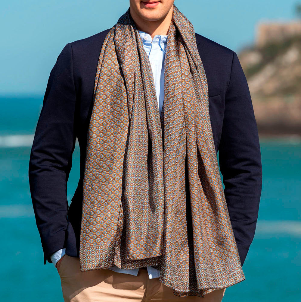 Man wearing a sophisticated brown silk scarf with an elegant geometric pattern in earthy tones, draped over a navy suit by the seaside.