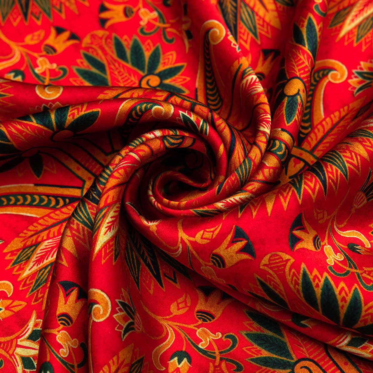 Close-up of a red silk scarf with golden and green floral motifs, neatly rolled into a spiral to highlight its lustrous texture and intricate design.