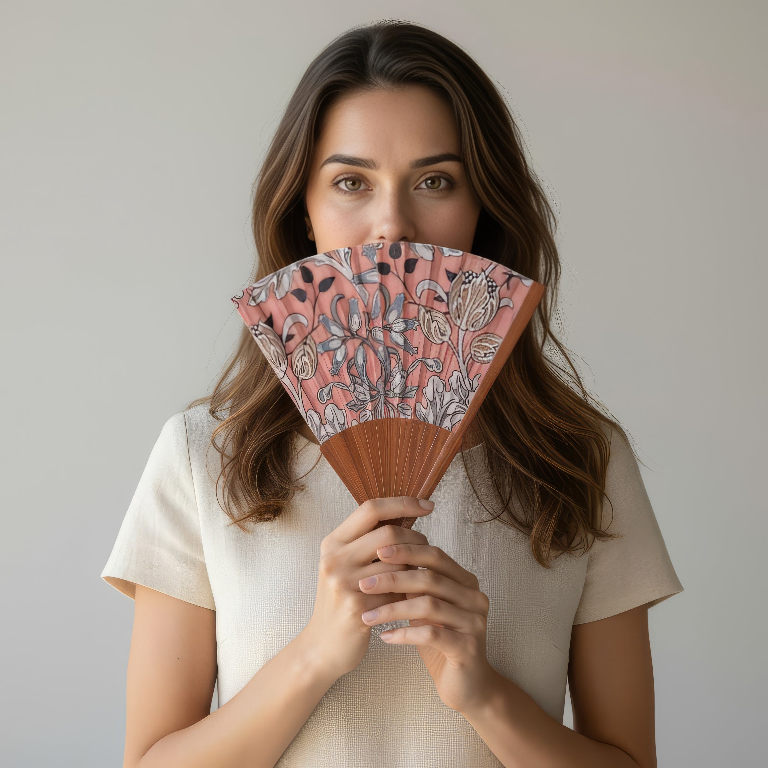 A woman with long brown hair holds the Folding Silk Fan with Floral Print Elinor, partially covering her face and revealing only her eyes. She wears a short-sleeve light beige top against a plain, light background.