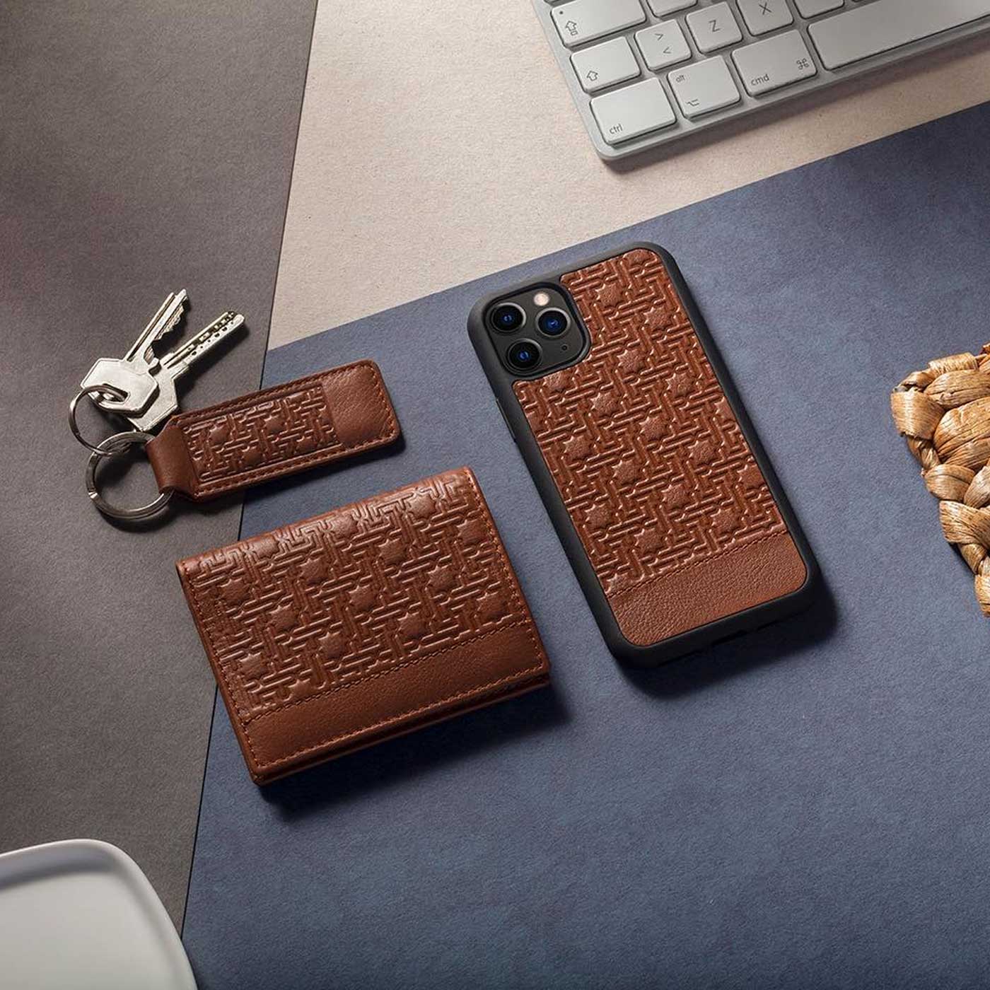 A set of brown leather accessories with a woven pattern, including a keychain, wallet, and phone case, are arranged on a desk near keys, a keyboard, and a braided cord.
