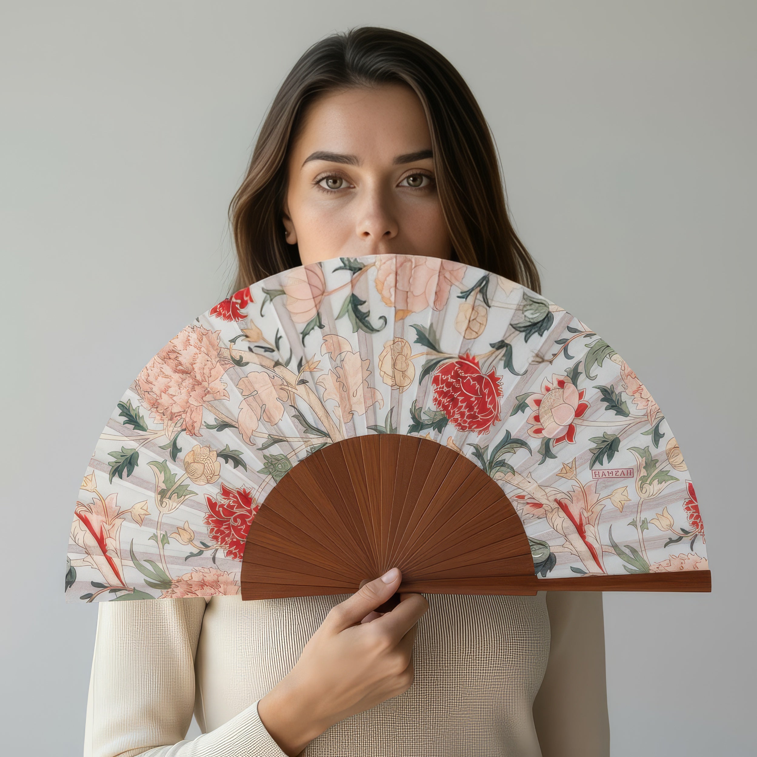 A woman with long brown hair holds the Floral Design Fan William Morris Print Odette, partially covering her face against a plain neutral background.
