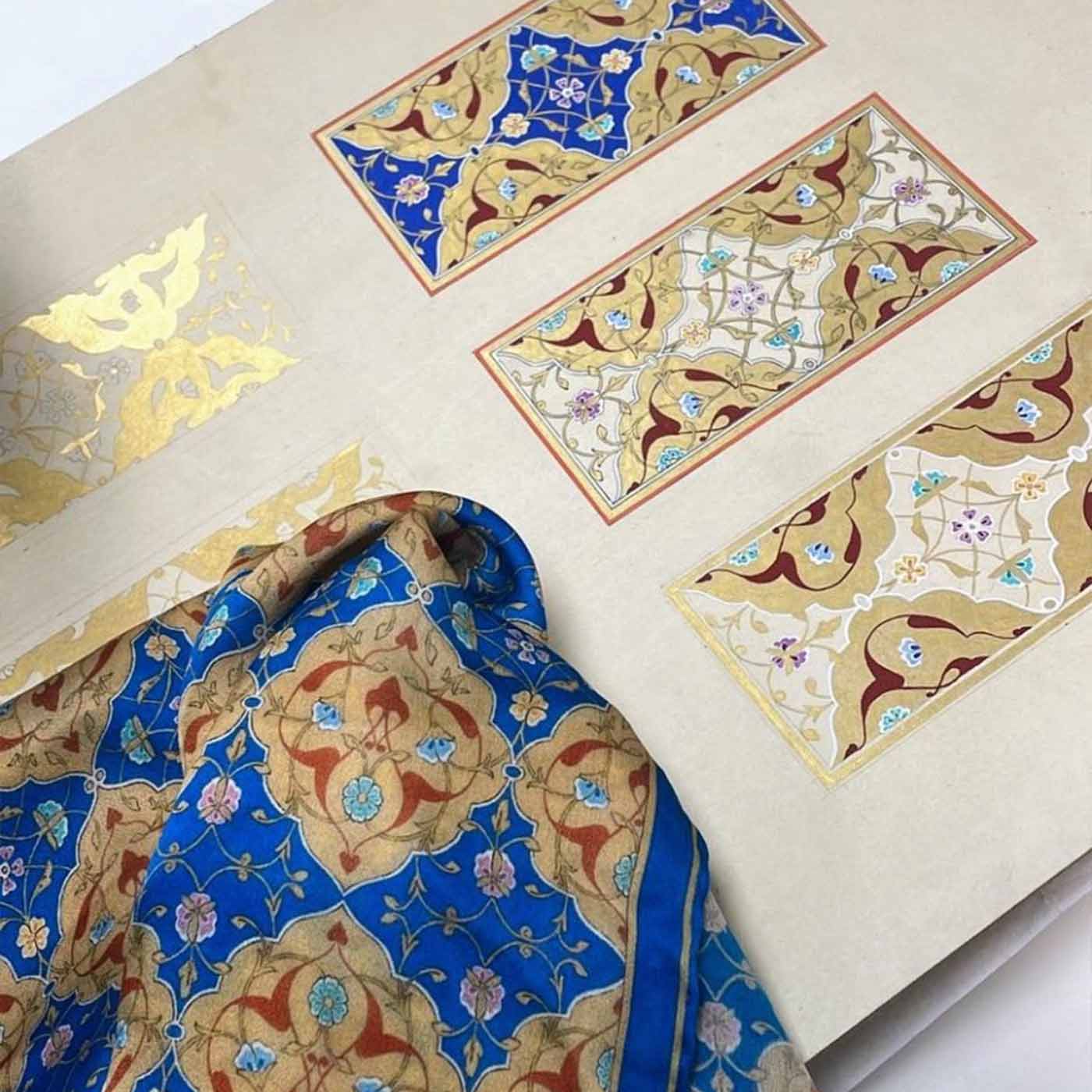 A patterned blue and gold fabric is draped over a page displaying intricate, rectangular Islamic geometric designs in blue, gold, red, and cream tones, with one design featuring gold leaf accents.