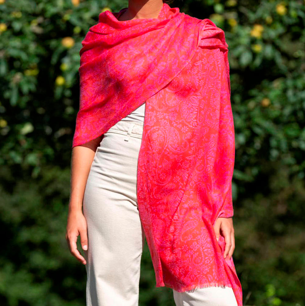 Pink wool and silk scarf with intricate paisley patterns, draped elegantly over her shoulders. The luxurious fabric catches the sunlight, highlighting its rich texture and soft drape.