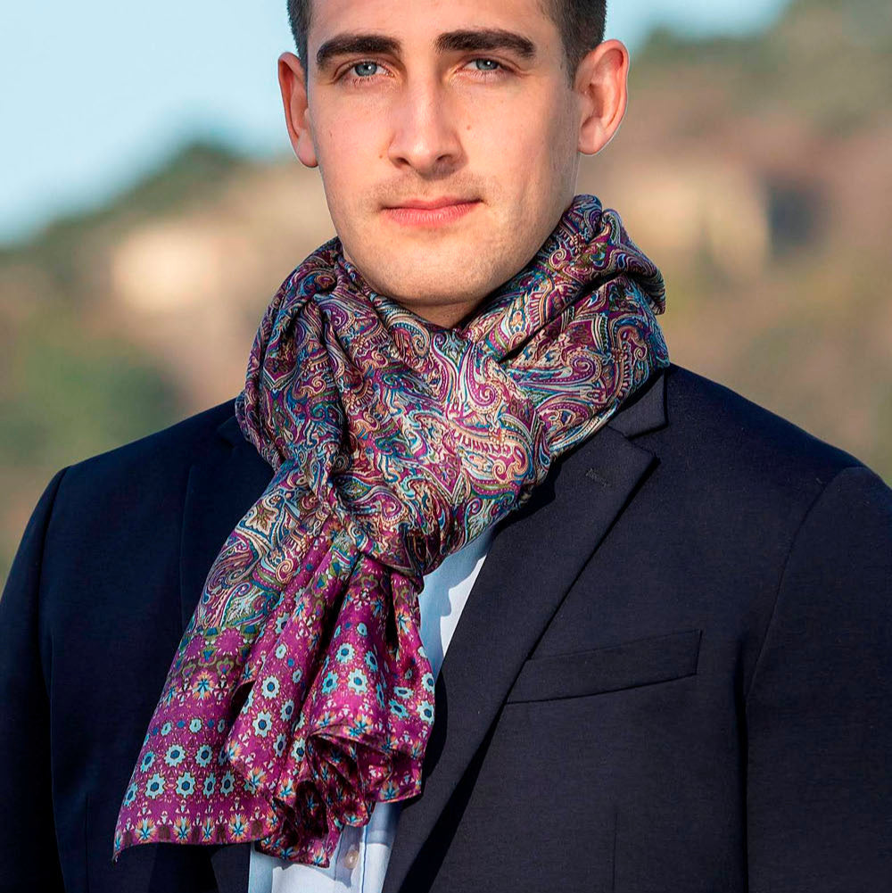 Man wearing an elegant purple silk scarf with intricate paisley patterns in shades of purple and blue, tied stylishly over a suit for a sophisticated look.