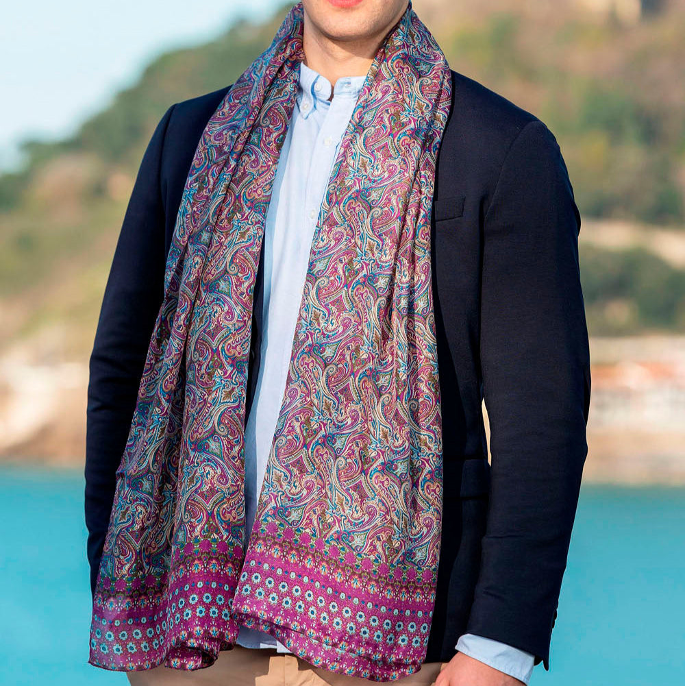 Man wearing a luxurious purple silk scarf with intricate paisley patterns in shades of purple and blue, elegantly draped over a suit by the seaside.