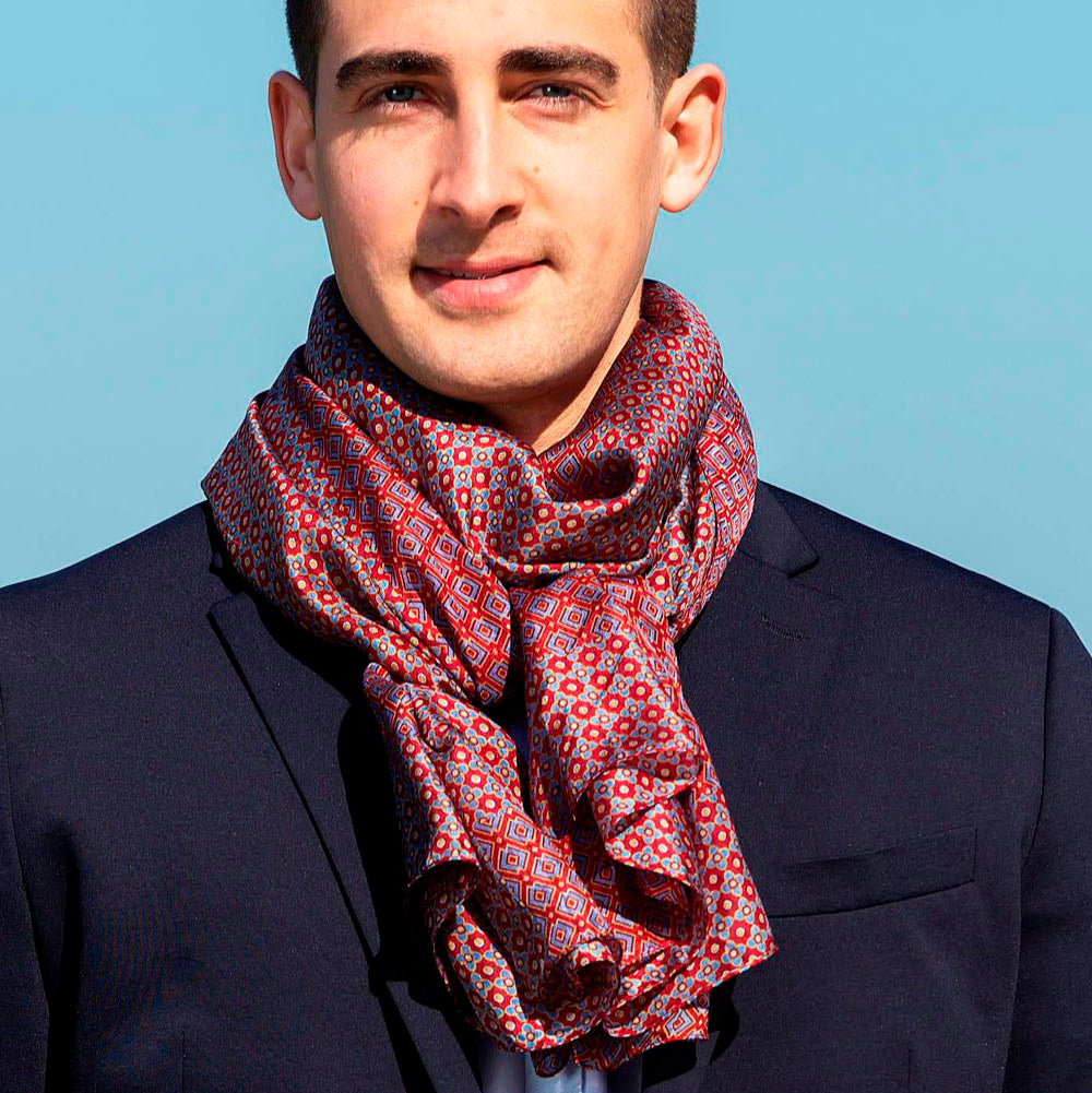 Man in a navy blazer wearing a red silk scarf with intricate geometric patterns, tied in a stylish knot. The bright blue sky enhances the luxurious and elegant look.