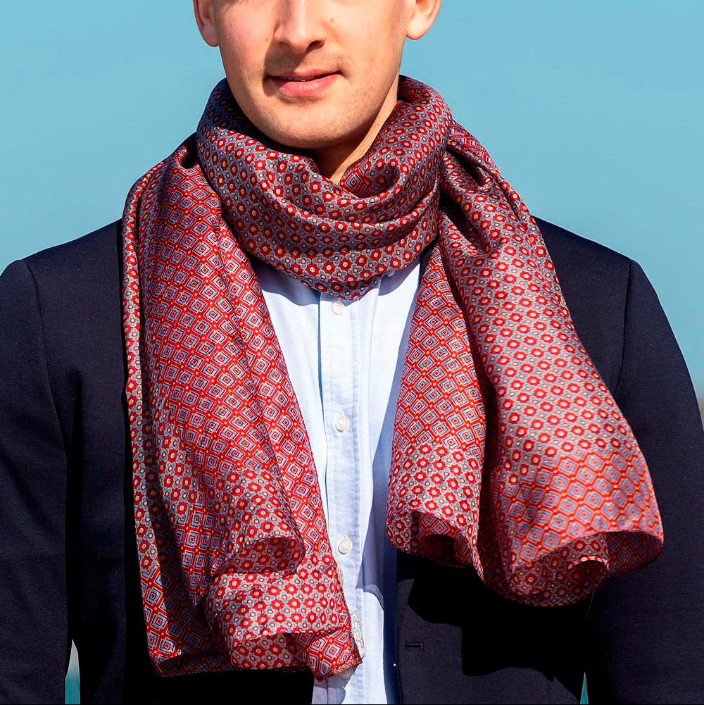 Man wearing a red silk scarf with a geometric pattern, wrapped elegantly around his neck, paired with a navy blazer and white shirt, with a clear blue sky in the background.