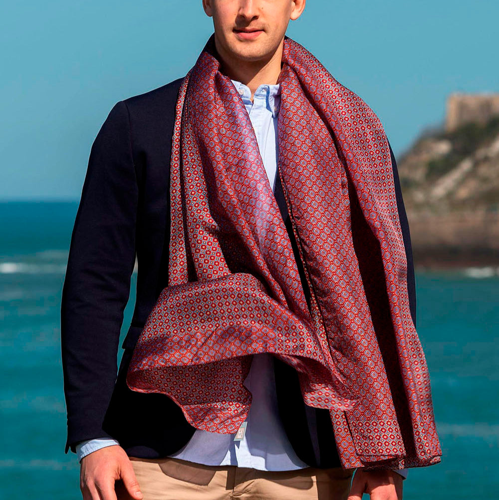 Man wearing a luxurious red silk scarf with an elegant geometric pattern, draped over a navy suit, standing against a scenic seaside backdrop.