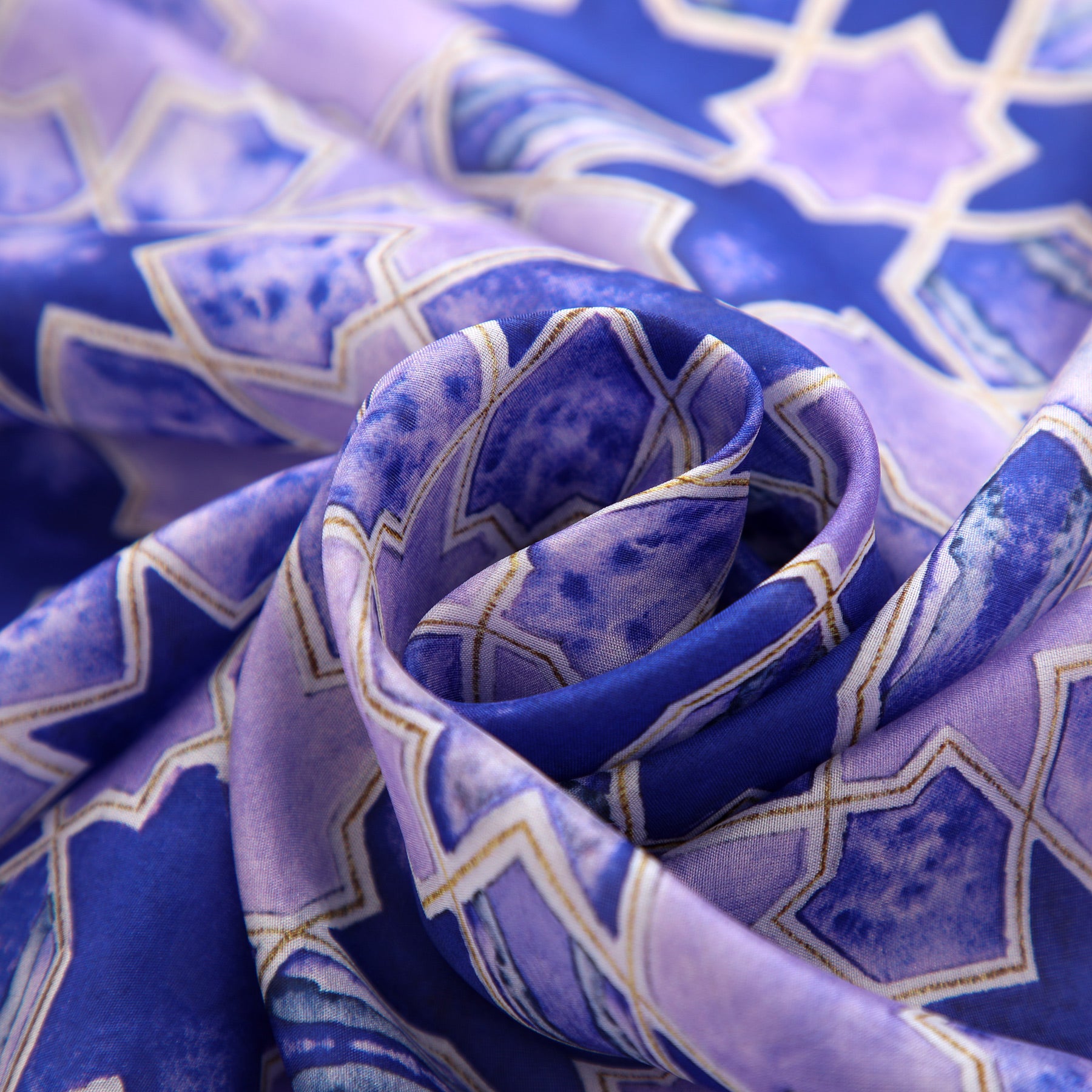 A close-up of the Purple Silk Scarf with Islamic Art Print Sevilla, showcasing a blue and purple geometric pattern inspired by Andalusian art with gold-outlined star and tile shapes, softly gathered in a spiral fold.