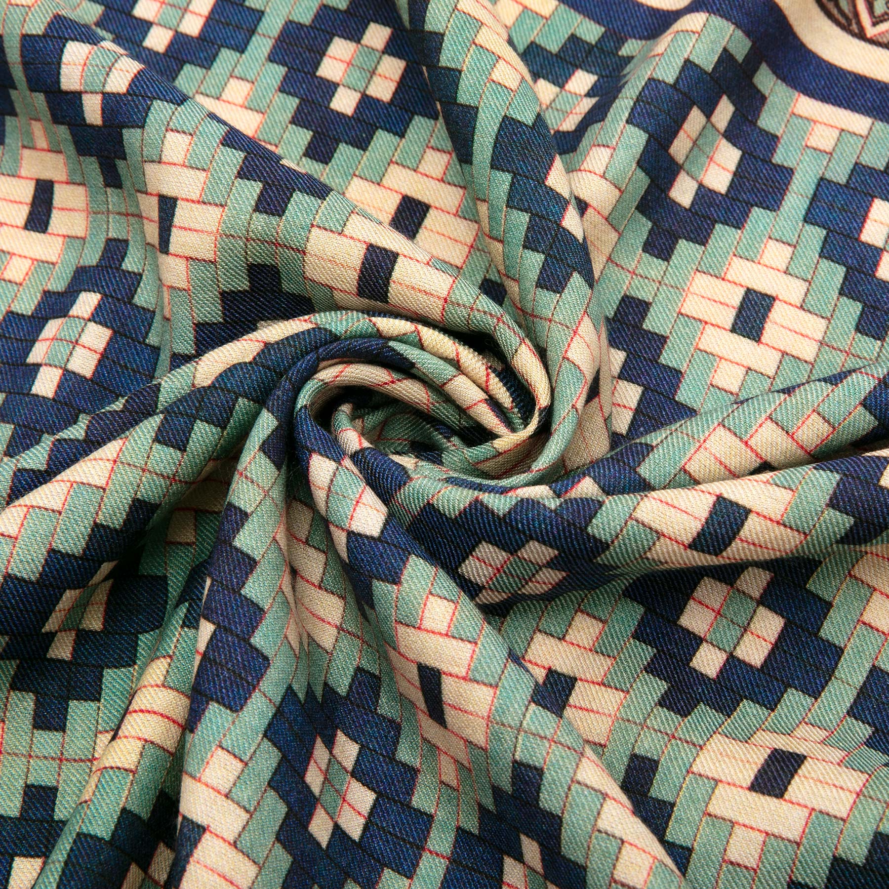 The Silk and Wool Scarf Ziyarat by Hamzah features a close-up of a multicolor geometric print with interlocking rectangles and diamonds in teal, navy, and cream. The luxurious fabric forms a spiral of folds showcasing its silk and wool blend design.